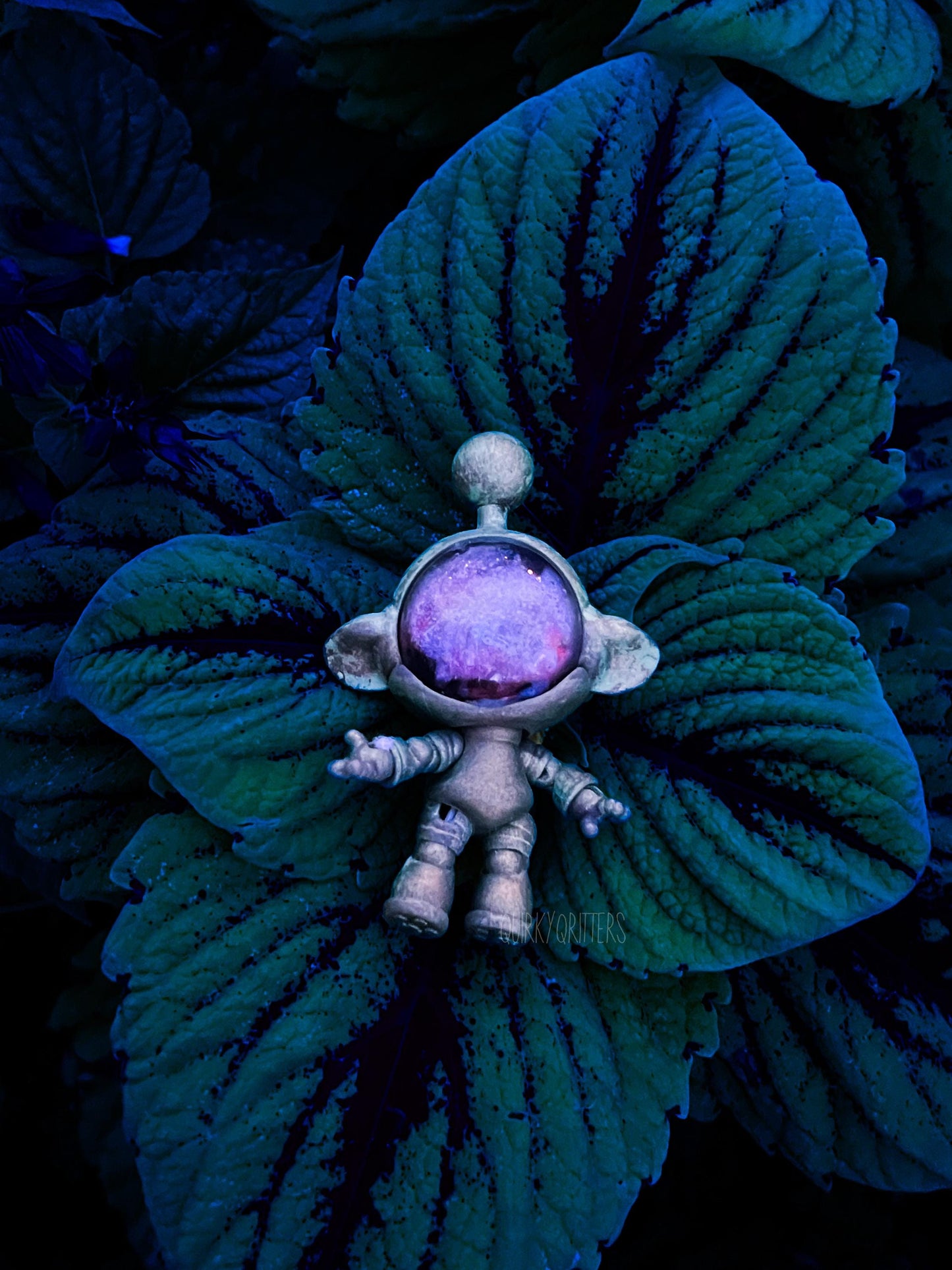 May include: A small, silver-colored astronaut figurine stands on a large, dark green leaf. The astronaut has a round, purple-tinted visor and outstretched arms. The background is filled with more of the dark green leaves, creating a contrasting backdrop. The figurine has the text 'kyorifters' on it.