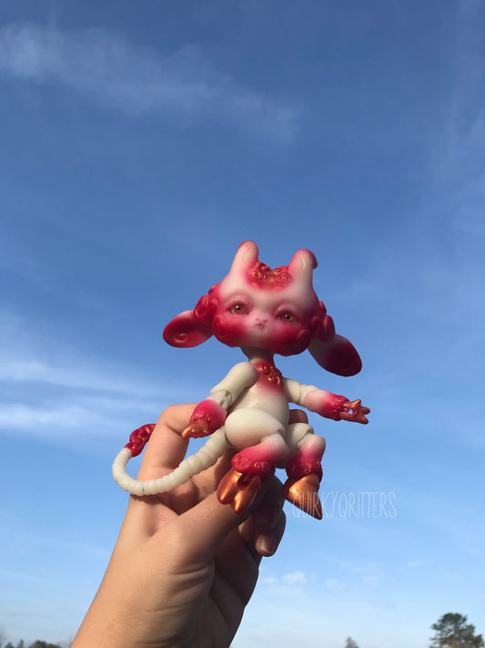 May include: A small, white and red, articulated doll with a long, curled tail. The doll has a human-like face with large, round eyes and a small, pointed nose. The doll is wearing a gold-colored, metallic paint on its feet and hands. The doll is posed in a sitting position with its arms outstretched.