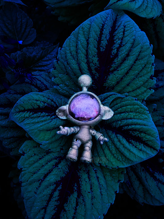 May include: A small, silver-colored astronaut figurine stands on a large, dark green leaf. The astronaut has a round, purple-tinted visor and outstretched arms. The background is filled with more of the dark green leaves, creating a contrasting backdrop. The figurine has the text &#39;kyorifters&#39; on it.