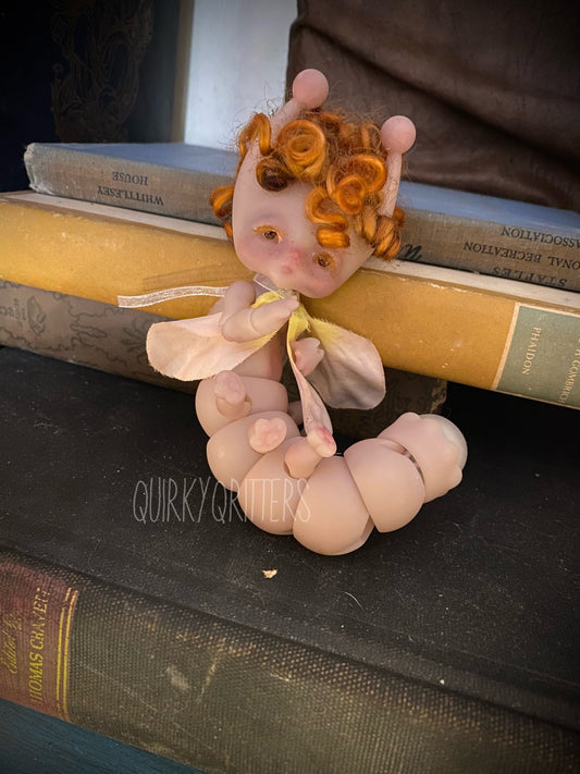 May include: A pink and white segmented caterpillar doll with a yellow shirt and orange curly hair. The doll is sitting on a stack of books. The text &#39;QUIRKYCRITTERS&#39; is visible on the bottom book.